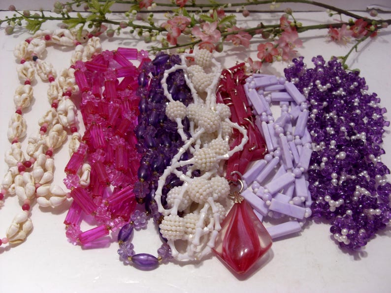 May include: A collection of six necklaces featuring various designs: a white seashell strand, a bright pink rectangular bead strand, a deep purple bead strand, a white textured bead strand, a light lavender rectangular bead strand, and a purple beaded strand with a large red diamond-shaped pendant.