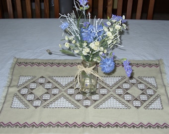 May include: A beige table runner with a geometric pattern in brown and pink. A glass vase with a bouquet of blue and white flowers sits on the runner.