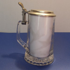 May include: Clear glass beer stein with a silver-colored metal lid and handle. The glass has a decorative etched design. The stein is sitting on a blue surface.