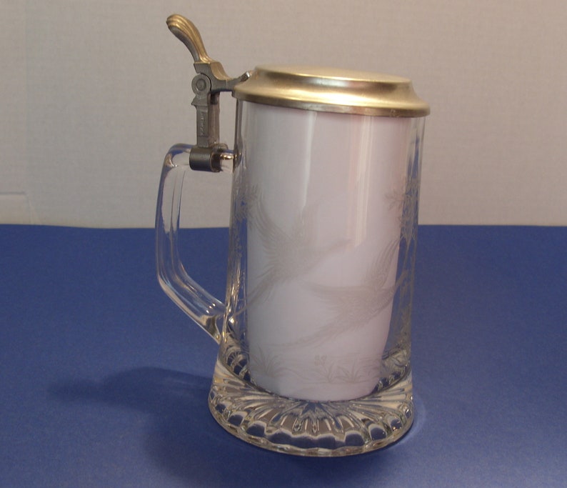 May include: Clear glass beer stein with a silver-colored metal lid and handle. The glass has a decorative etched design. The stein is sitting on a blue surface.