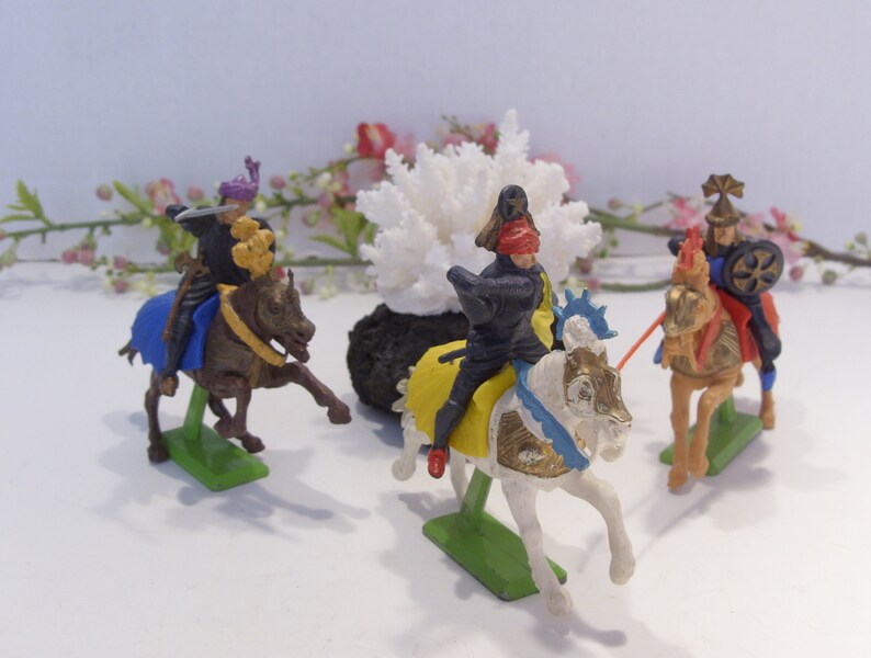 May include: Four plastic toy knights on horseback, each with a different colored horse and armor. The knights are posed in a battle scene, with swords drawn and shields raised. The horses are all in motion, with their legs raised in mid-stride.