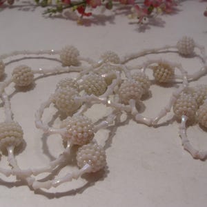 May include: A long necklace featuring a series of white, spherical, textured beaded clusters alternating with smooth, white, elongated, bone-shaped spacer beads. The necklace is arranged in a loose, tangled loop, showcasing the repeating pattern of the round, bumpy beads and the slender, curved connectors throughout the entire length of the jewelry piece.
