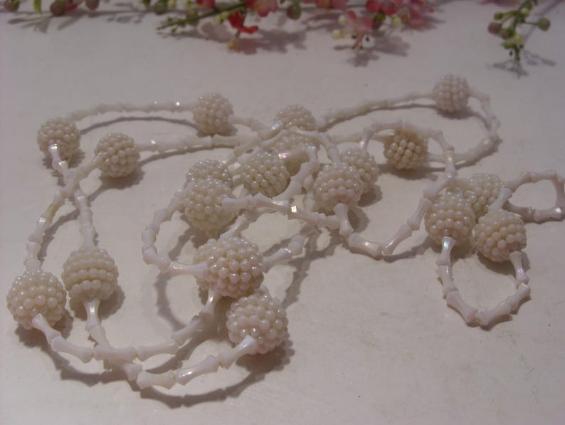 May include: A long necklace featuring a series of white, spherical, textured beaded clusters alternating with smooth, white, elongated, bone-shaped spacer beads. The necklace is arranged in a loose, tangled loop, showcasing the repeating pattern of the round, bumpy beads and the slender, curved connectors throughout the entire length of the jewelry piece.