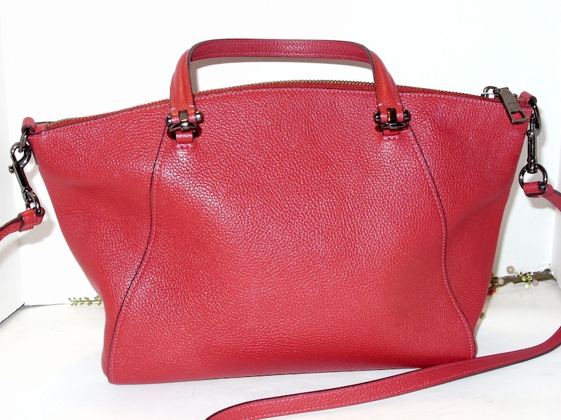 May include: A red leather handbag with a top handle and a detachable shoulder strap. The bag has a zip closure and dark metal hardware. The leather has a textured grain.