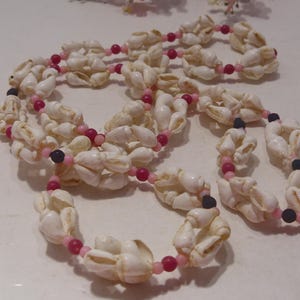 May include: A long necklace featuring clusters of white cowrie shells interspersed with small, round beads in shades of bright pink, light pink, and black. The shells are arranged in repeating segments along the length of the strand.