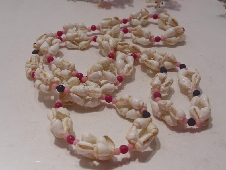May include: A long necklace featuring clusters of white cowrie shells interspersed with small, round beads in shades of bright pink, light pink, and black. The shells are arranged in repeating segments along the length of the strand.