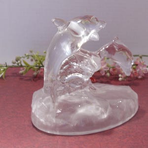 May include: A clear glass dolphin sculpture with two dolphins leaping from the water. The sculpture is on a maroon surface with pink and green floral accents in the background. The dolphins are detailed and appear to be in motion.