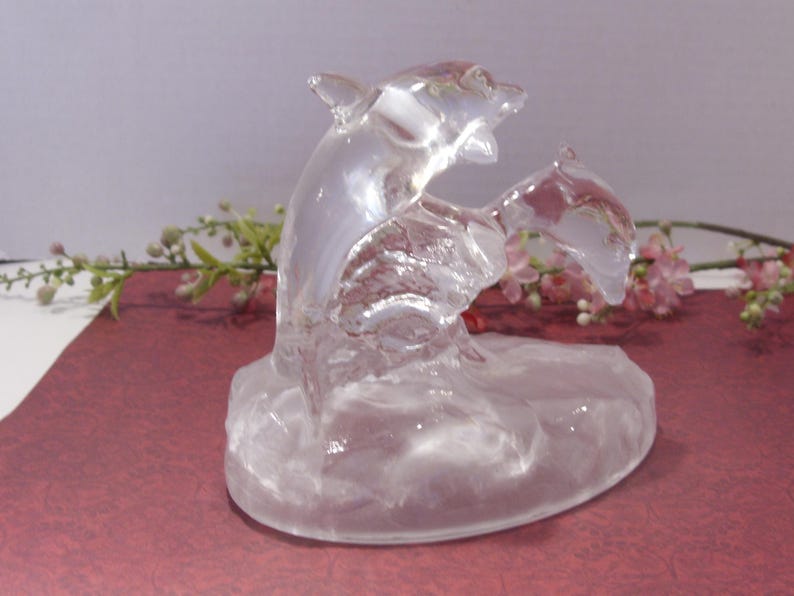 May include: A clear glass dolphin sculpture with two dolphins leaping from the water. The sculpture is on a maroon surface with pink and green floral accents in the background. The dolphins are detailed and appear to be in motion.