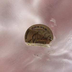 May include: A close-up of a light pink, possibly glass, surface with a partially torn, circular sticker. The sticker is gold with black text that reads "Plus de 24% de" and "Cristal d'Arques".