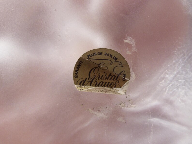 May include: A close-up of a light pink, possibly glass, surface with a partially torn, circular sticker. The sticker is gold with black text that reads "Plus de 24% de" and "Cristal d'Arques".