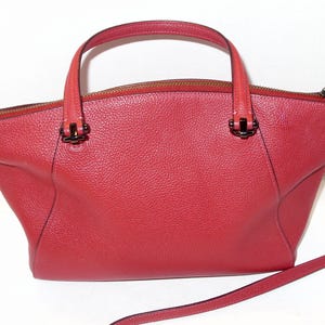 May include: A red leather handbag with a top handle and a detachable shoulder strap. The bag has a zipper closure and metal hardware. The bag's design is a trapezoid shape, with a textured surface.