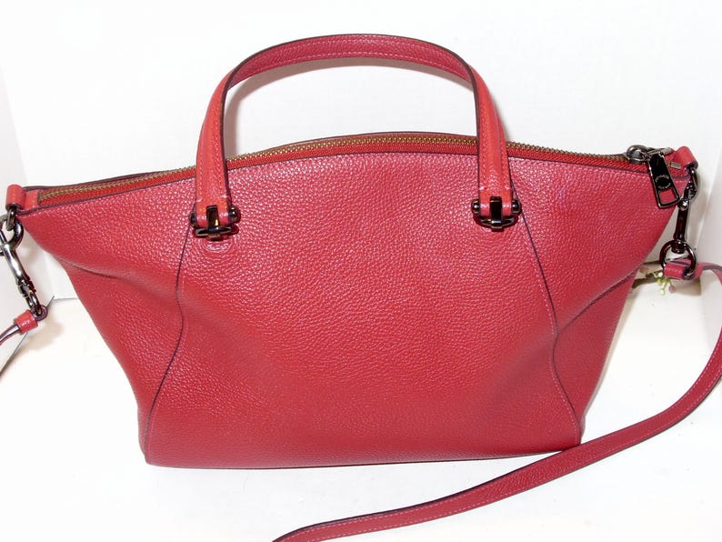 May include: A red leather handbag with a top handle and a detachable shoulder strap. The bag has a zipper closure and metal hardware. The bag's design is a trapezoid shape, with a textured surface.