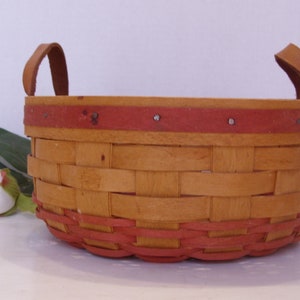 May include: A woven basket with two leather handles. The basket is made of natural wood and has a red painted rim.