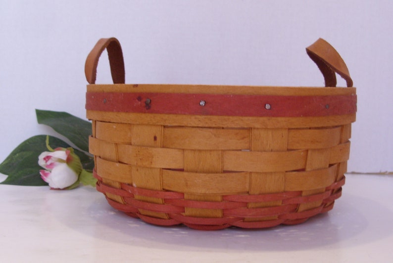 May include: A woven basket with two leather handles. The basket is made of natural wood and has a red painted rim.