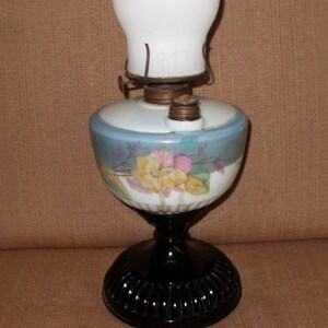 May include: A vintage oil lamp with a white glass shade and a black base. The lamp features a floral design with yellow, pink, and blue flowers.