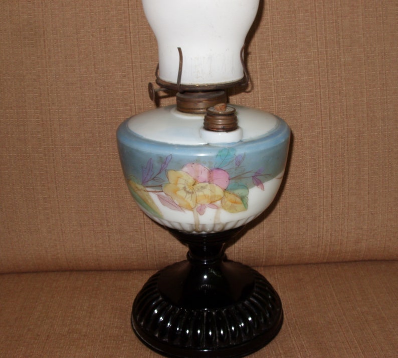 May include: A vintage oil lamp with a white glass shade and a black base. The lamp features a floral design with yellow, pink, and blue flowers.