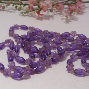 May include: A long necklace featuring translucent purple beads in alternating shapes of elongated ovals and small five-petaled flowers. The beads are strung together on a thin cord, creating a continuous strand of violet-colored floral and oval elements.