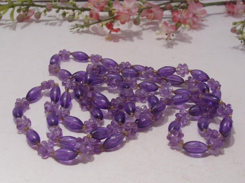May include: A long necklace featuring translucent purple beads in alternating shapes of elongated ovals and small five-petaled flowers. The beads are strung together on a thin cord, creating a continuous strand of violet-colored floral and oval elements.