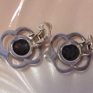 May include: A pair of silver clip-on earrings with a floral design and black stones in the center.