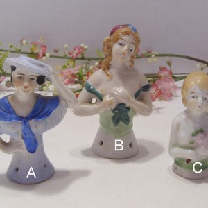 May include: Three antique porcelain half dolls. One doll is dressed as a sailor, another has a green bodice, and the third holds a pink flower. Each doll has a hole at the base.