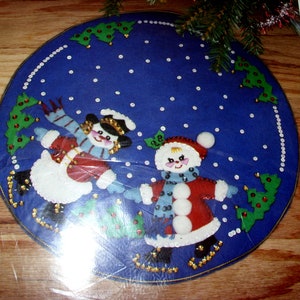 May include: A blue felt Christmas tree skirt featuring two snowmen ice skating. The snowmen wear red coats, hats, and scarves, with white snow and green trees around the border. The skirt is round.