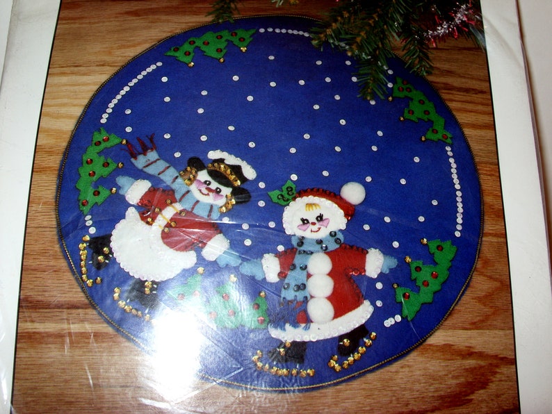 May include: A blue felt Christmas tree skirt featuring two snowmen ice skating. The snowmen wear red coats, hats, and scarves, with white snow and green trees around the border. The skirt is round.