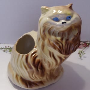 May include: A vintage ceramic cat planter in a seated position. The cat has a cream-colored body with brown fur accents and blue eyes. The planter has a round opening on its back for plants or flowers.