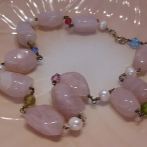 May include: A delicate bracelet featuring large, polished rose quartz beads, interspersed with small white pearls and colorful accent beads. The bracelet is strung on a delicate chain, creating a soft, feminine aesthetic.