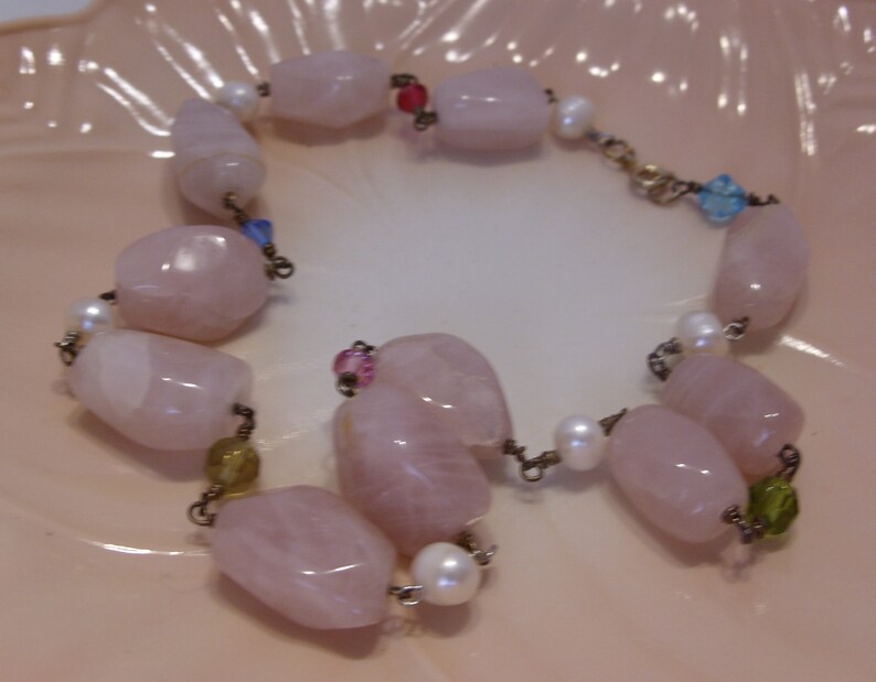 May include: A delicate bracelet featuring large, polished rose quartz beads, interspersed with small white pearls and colorful accent beads. The bracelet is strung on a delicate chain, creating a soft, feminine aesthetic.