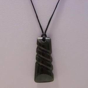 May include: A dark green pendant necklace with a carved spiral design. The pendant is rectangular with rounded edges and is suspended from a black cord. The necklace is displayed against a light background.