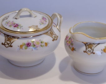 May include: A white porcelain sugar bowl with a lid and a creamer. Both pieces are decorated with pink, yellow, and purple floral patterns and gold trim. The sugar bowl has a handle and the creamer has a spout.