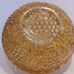 Puede incluir: Un cuenco de vidrio &aacute;mbar iridiscente con un dise&ntilde;o de uvas y hojas. El cuenco tiene forma redondeada con un dise&ntilde;o en relieve detallado. El vidrio tiene una superficie brillante y reflectante, probablemente vintage.