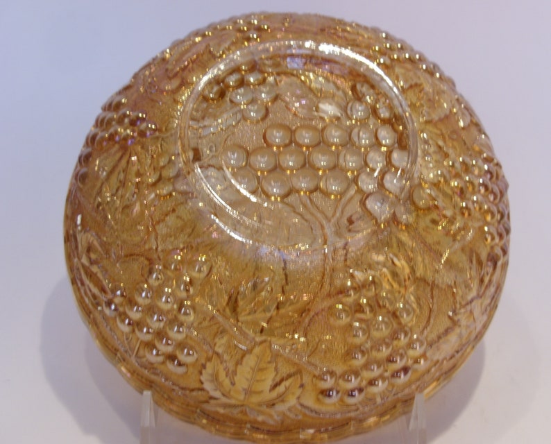 Puede incluir: Un cuenco de vidrio &aacute;mbar iridiscente con un dise&ntilde;o de uvas y hojas. El cuenco tiene forma redondeada con un dise&ntilde;o en relieve detallado. El vidrio tiene una superficie brillante y reflectante, probablemente vintage.