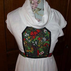 May include: White dress with a cowl neck and short sleeves. The bodice features a floral panel with red, yellow, and white flowers on a black background. A matching floral belt accents the waist, creating a vintage-inspired look.