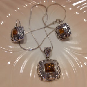 K&ouml;nnte beinhalten: Ein silbernes Halsketten- und Ohrring-Set mit einem braunen Edelstein in jedem St&uuml;ck. Die Halskette hat eine zarte Kette und einen Anh&auml;nger mit einem verzierten Design. Die Ohrringe sind Ohrh&auml;nger mit einem &auml;hnlichen Design.