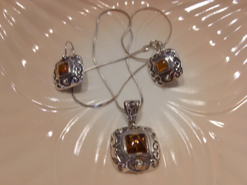 K&ouml;nnte beinhalten: Ein silbernes Halsketten- und Ohrring-Set mit einem braunen Edelstein in jedem St&uuml;ck. Die Halskette hat eine zarte Kette und einen Anh&auml;nger mit einem verzierten Design. Die Ohrringe sind Ohrh&auml;nger mit einem &auml;hnlichen Design.