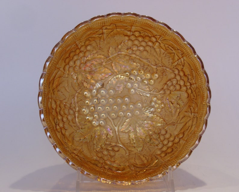 Puede incluir: Un cuenco de vidrio &aacute;mbar iridiscente con borde ondulado. El cuenco presenta un dise&ntilde;o en relieve detallado de uvas y hojas. El vidrio tiene una calidad brillante y reflectante. El cuenco est&aacute; sobre un fondo neutro.