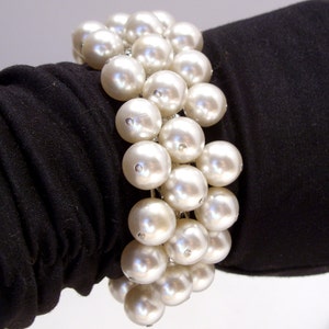 May include: A cluster bracelet featuring large, round, faux pearls. The pearls are a creamy white color and are strung together with silver-toned metal. The bracelet is shown against a black background.