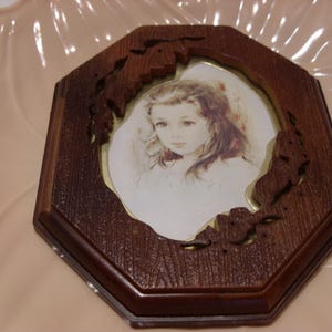 May include: An octagonal wooden picture frame with a portrait of a young girl. The frame is dark brown with carved floral accents. The portrait features a girl with long, wavy hair and a soft, neutral color palette.