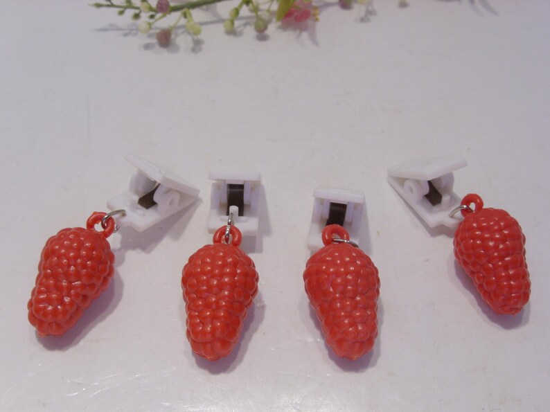 Op de afbeelding: Vier witte plastic clips met rode aardbei bedels. De bedels zijn felrood van kleur en hebben een gestructureerd oppervlak, dat op de vrucht lijkt. De clips zijn gerangschikt op een wit oppervlak.