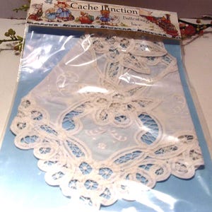 May include: A piece of off-white Battenburg lace featuring intricate floral and scrollwork patterns with openwork mesh details. The lace is sealed inside a clear plastic package with a header card labeled Cache Junction and Battenburg lace.