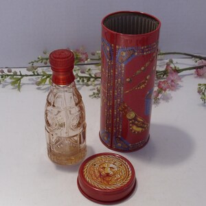 May include: A clear glass perfume bottle with a red cap stands next to a cylindrical red metal container. The container features intricate gold and blue patterns, and its separate lid displays a detailed embossed lion head design in gold.