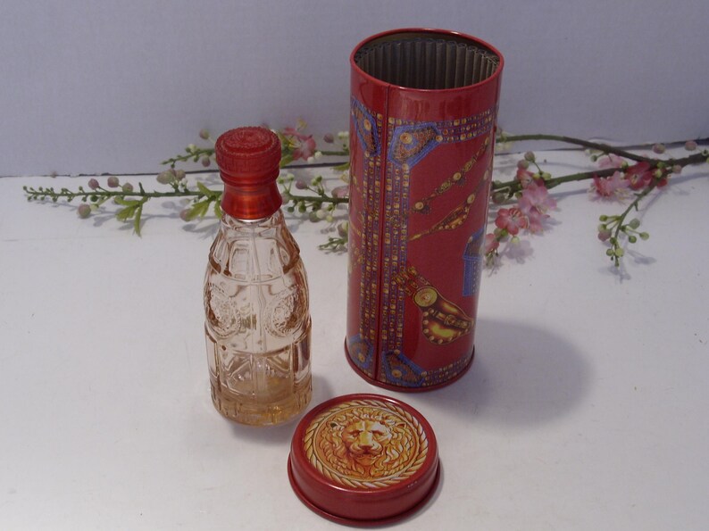 May include: A clear glass perfume bottle with a red cap stands next to a cylindrical red metal container. The container features intricate gold and blue patterns, and its separate lid displays a detailed embossed lion head design in gold.