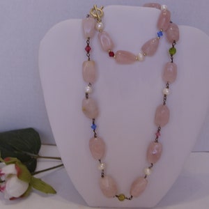 May include: A necklace featuring large, light pink, irregular-shaped beads interspersed with small white pearls and colorful accent beads. The necklace has a gold-colored clasp and is displayed on a white bust.