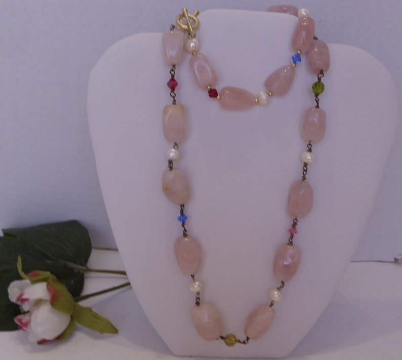 May include: A necklace featuring large, light pink, irregular-shaped beads interspersed with small white pearls and colorful accent beads. The necklace has a gold-colored clasp and is displayed on a white bust.