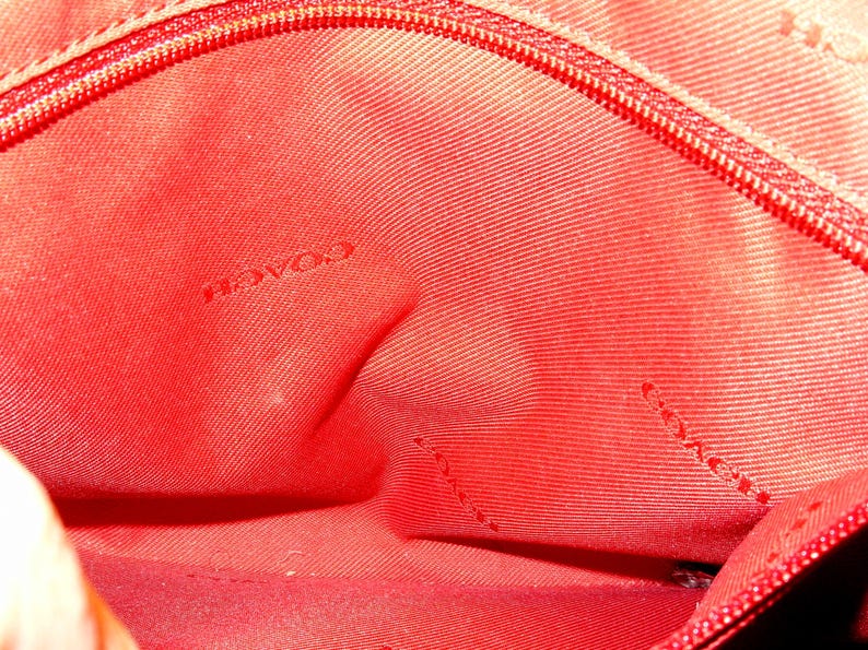 May include: Close-up of the interior of a red Coach handbag. The fabric is a solid red with a subtle texture. The word "COACH" is visible in the center of the image. A red zipper is visible along the top edge.