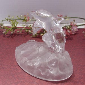 May include: A clear glass dolphin sculpture leaping from a textured base. The dolphin is detailed with fins and a curved body, appearing to jump through water. The sculpture is set against a burgundy background with floral accents.