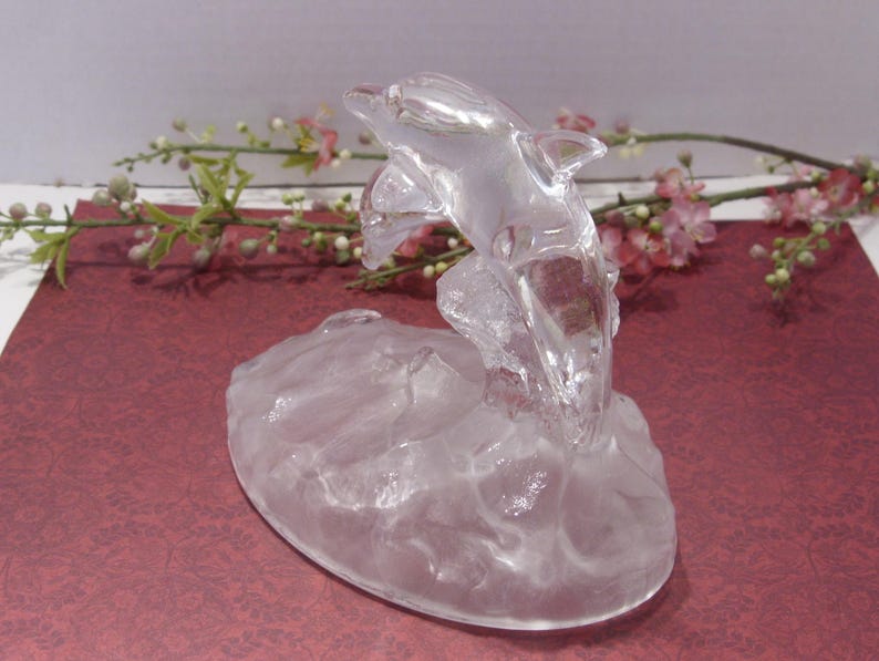 May include: A clear glass dolphin sculpture leaping from a textured base. The dolphin is detailed with fins and a curved body, appearing to jump through water. The sculpture is set against a burgundy background with floral accents.