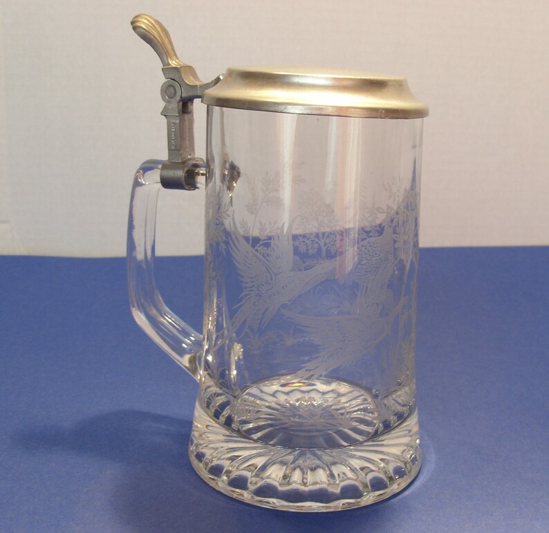 May include: Clear glass beer stein with a metal hinged lid and handle. The glass is etched with a design of birds and foliage. The base of the stein has a decorative, scalloped design.