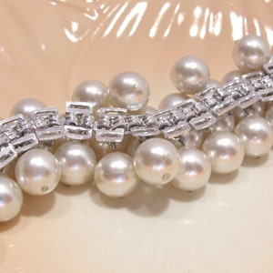 May include: A vintage bracelet featuring a silver-tone metal link design adorned with numerous large, round, creamy white faux pearls. The bracelet is displayed on a light peach-colored surface, showcasing its elegant design.